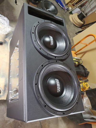Two large black subwoofers mounted in a custom black speaker enclosure. The enclosure has a rectangular design.