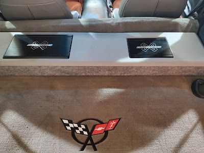 Car interior with silver panel holding two black amplifiers, over a carpet with a Corvette logo.