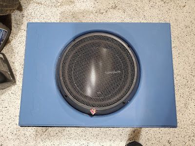 Blue rectangular subwoofer box with a black mesh-covered speaker in the center, placed on a concrete surface.