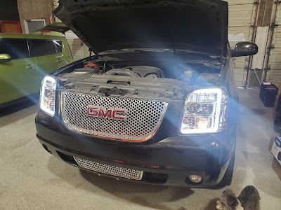 Black GMC Yukon with the hood open, showcasing custom bright white headlights and a chrome grille in a garage setting.