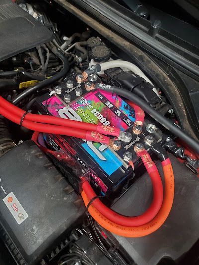 Car battery with red and orange cables in an engine compartment. The battery is colorful with a pink and blue design.