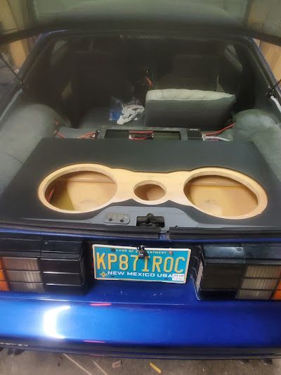 A blue car's trunk with a custom subwoofer enclosure. It has two large speaker openings, with a center cutout, and wood trim.