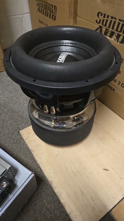 A large, black Sundown Audio subwoofer speaker with a chrome base sits on a wooden surface.