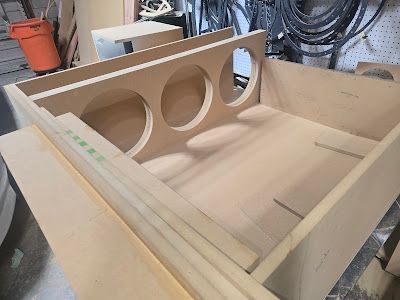 Wooden speaker box under construction, showing cutouts for three speaker drivers. Construction happening in a workshop.