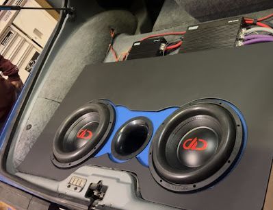Car trunk with a custom subwoofer setup: two black subwoofers flanking a blue port, mounted on a black panel.