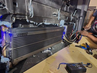 A car audio installer working on a vehicle, installing a large amplifier. The amplifier is silver with blue LED lights.