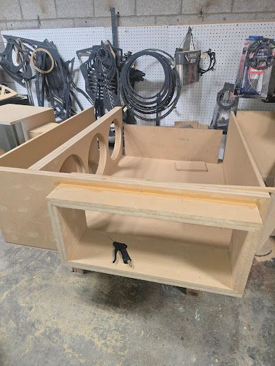 A custom-built wooden subwoofer enclosure with speaker holes and compartments in a workshop setting.