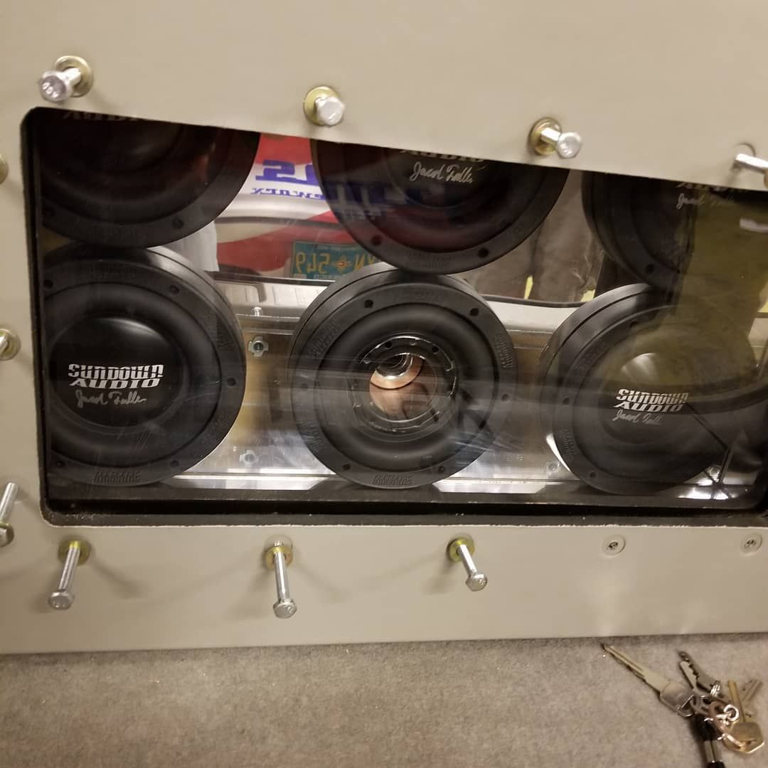 A rectangular speaker enclosure with six round black subwoofers visible behind a clear window, secured by bolts.