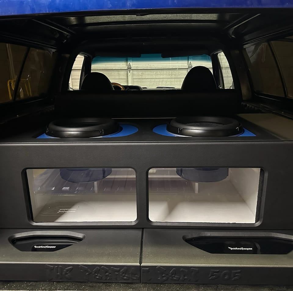 Custom car audio system with two subwoofers visible through plexiglass, mounted in the back of a truck with blue trim.