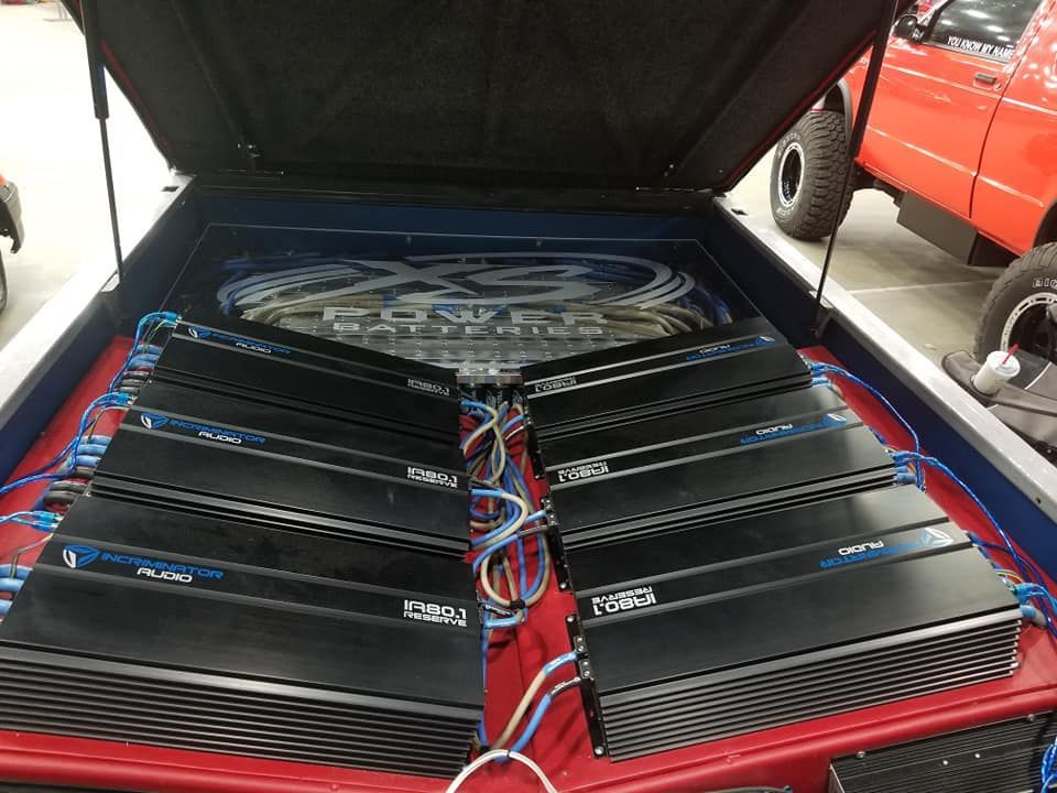 Car trunk with rows of black audio amplifiers, blue and white wiring, and custom artwork.
