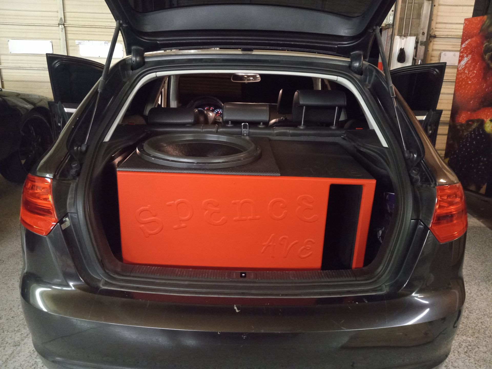 A black car's trunk with a large orange subwoofer box and speaker inside. The car's trunk is open in a garage setting.