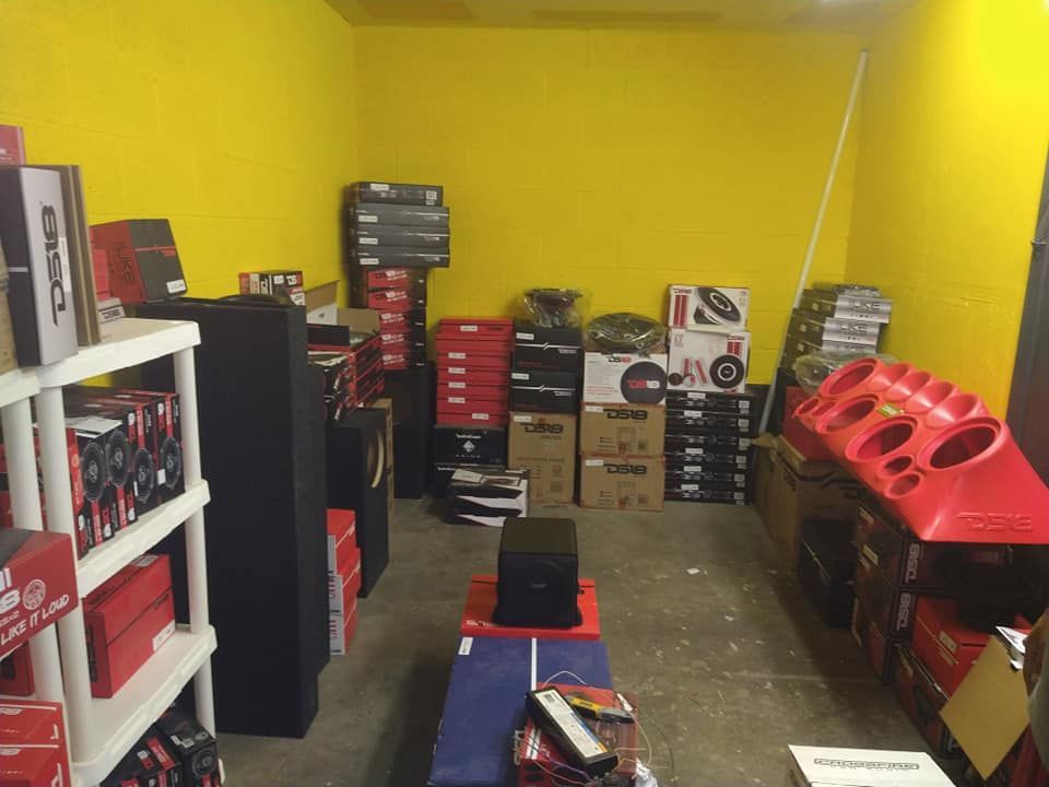 A cluttered room with yellow walls, showcasing car audio equipment like speakers and boxes, for sale.
