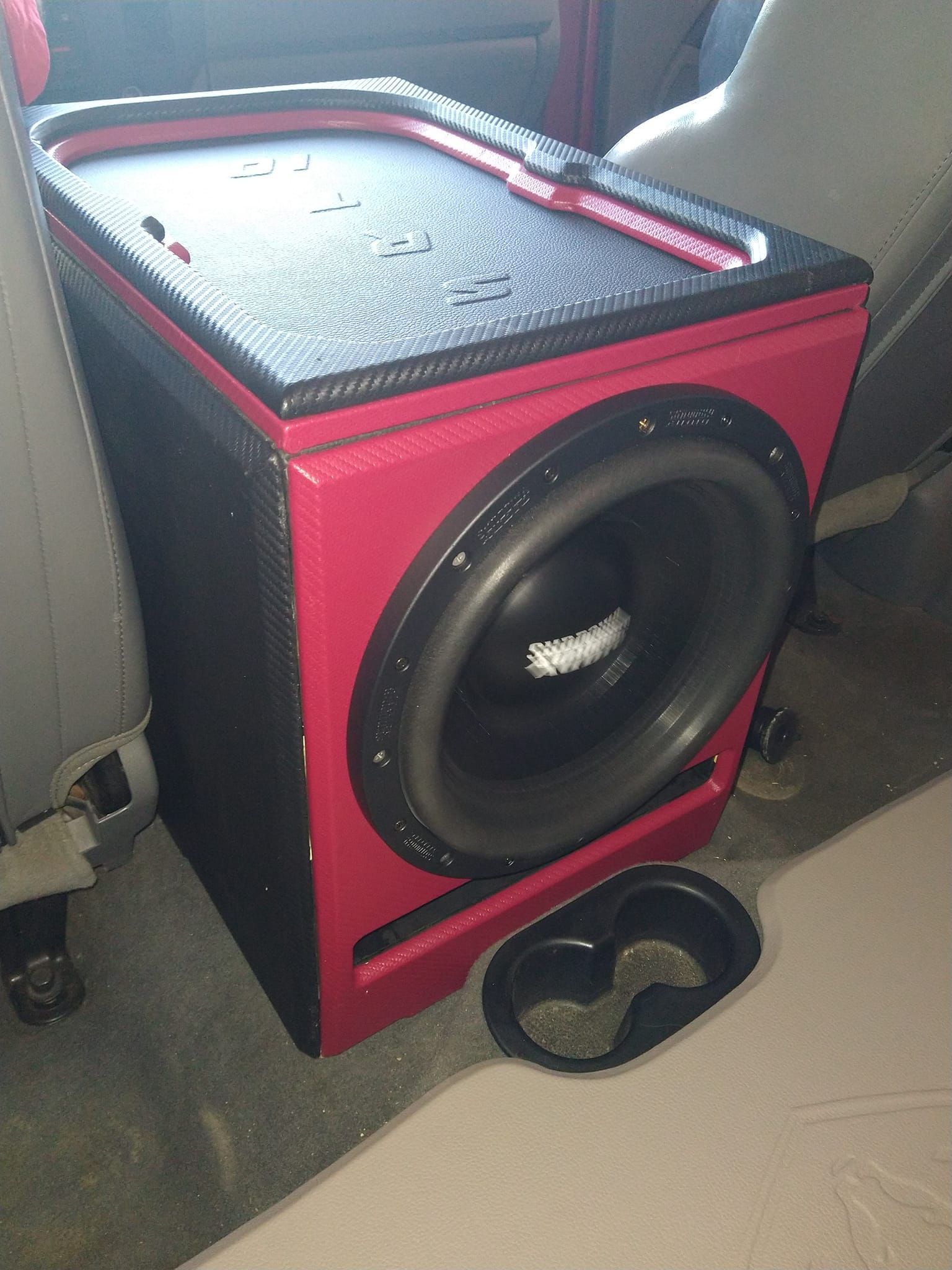 Red and black custom car audio subwoofer enclosure with a cup holder, placed in the back of a vehicle between the seats.