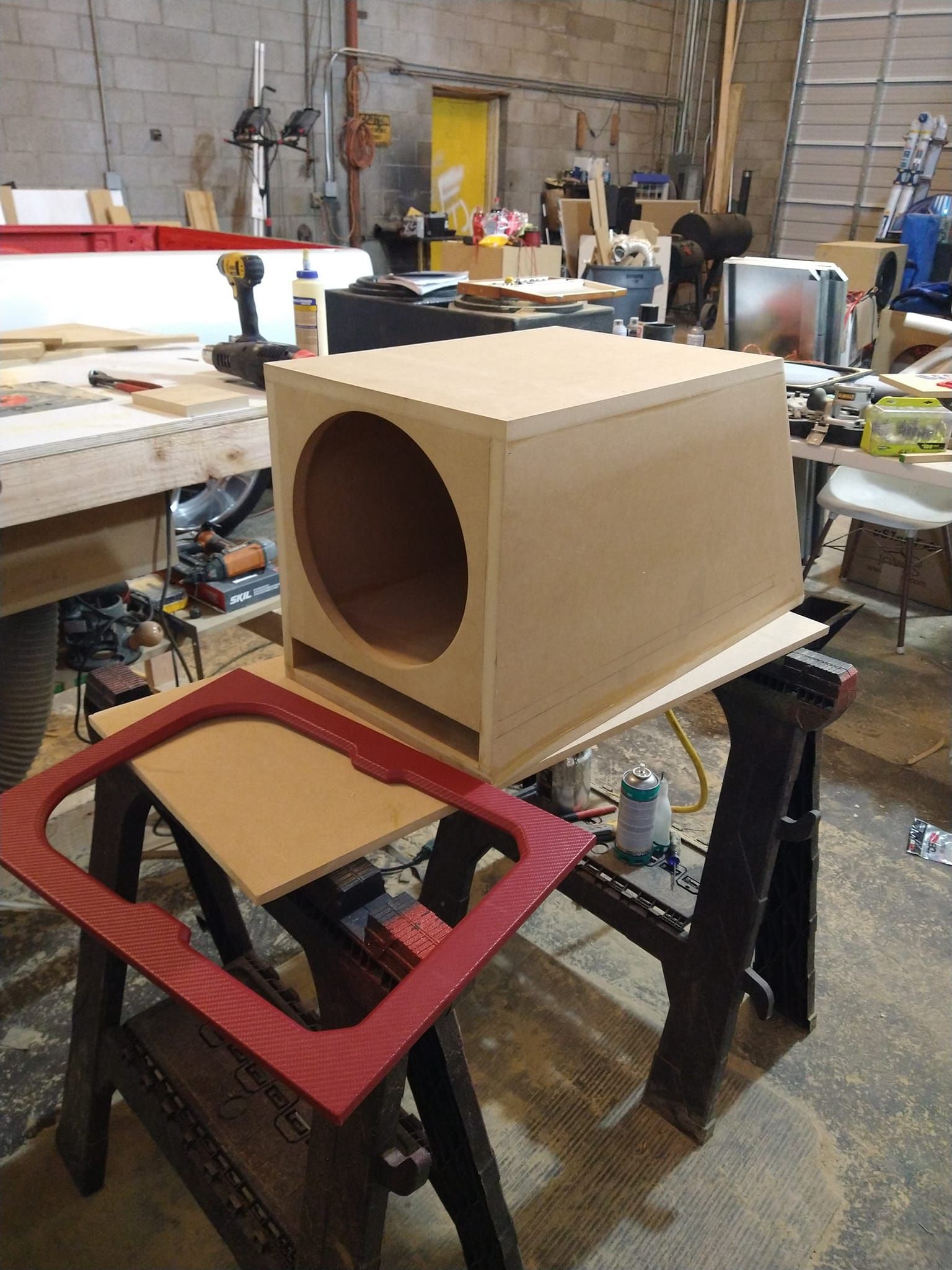 A wooden subwoofer box in a workshop, with a red speaker grille frame. The unfinished box rests on a sawhorse.