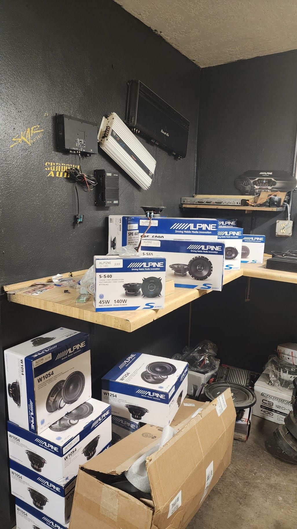 A cluttered workspace with car audio equipment: amplifiers, speakers, and boxes.
