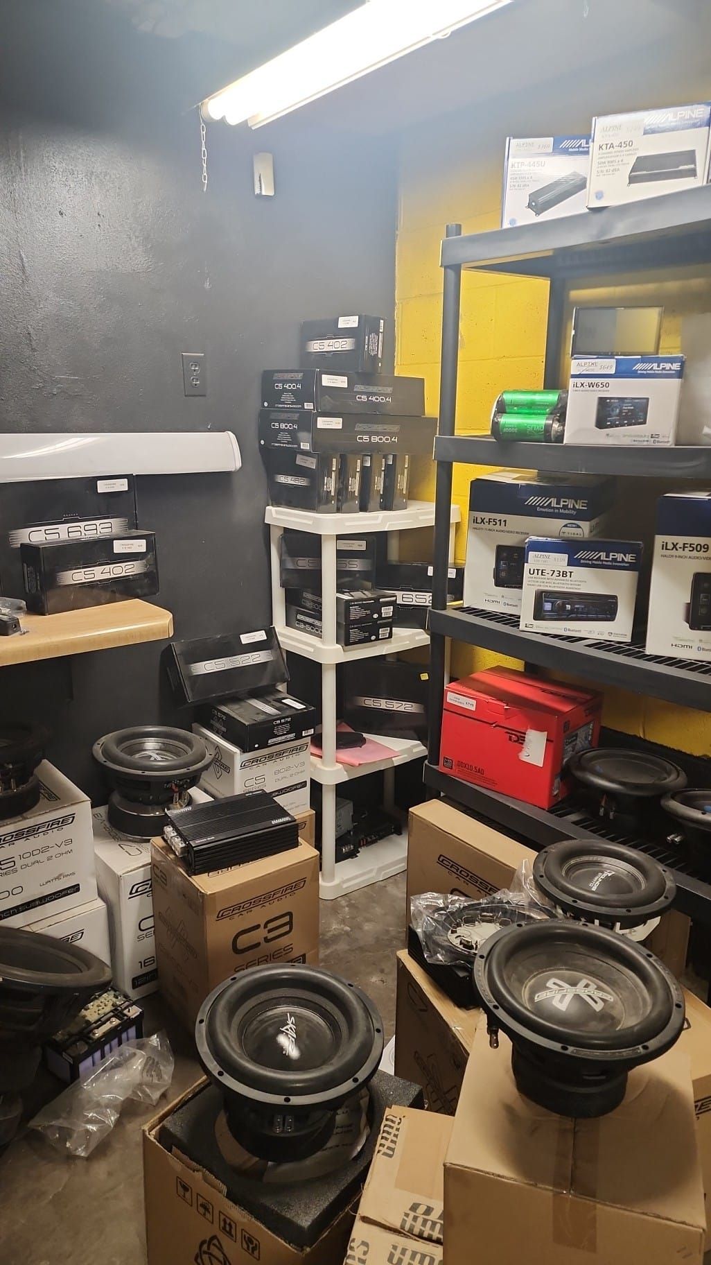 A storage room stocked with car audio equipment. Black subwoofers and amplifiers sit on boxes and shelves.