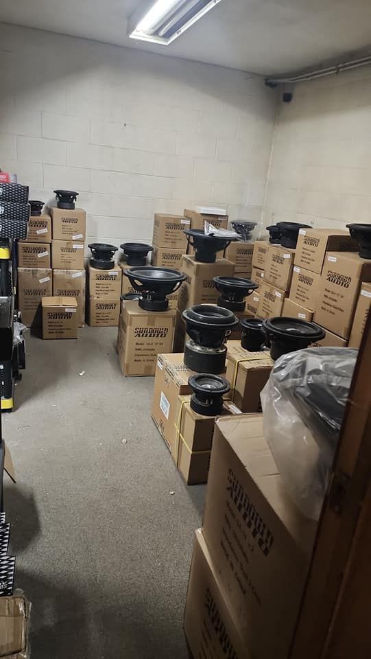 Warehouse filled with cardboard boxes, some with subwoofers on top. Overhead lighting illuminates the scene.