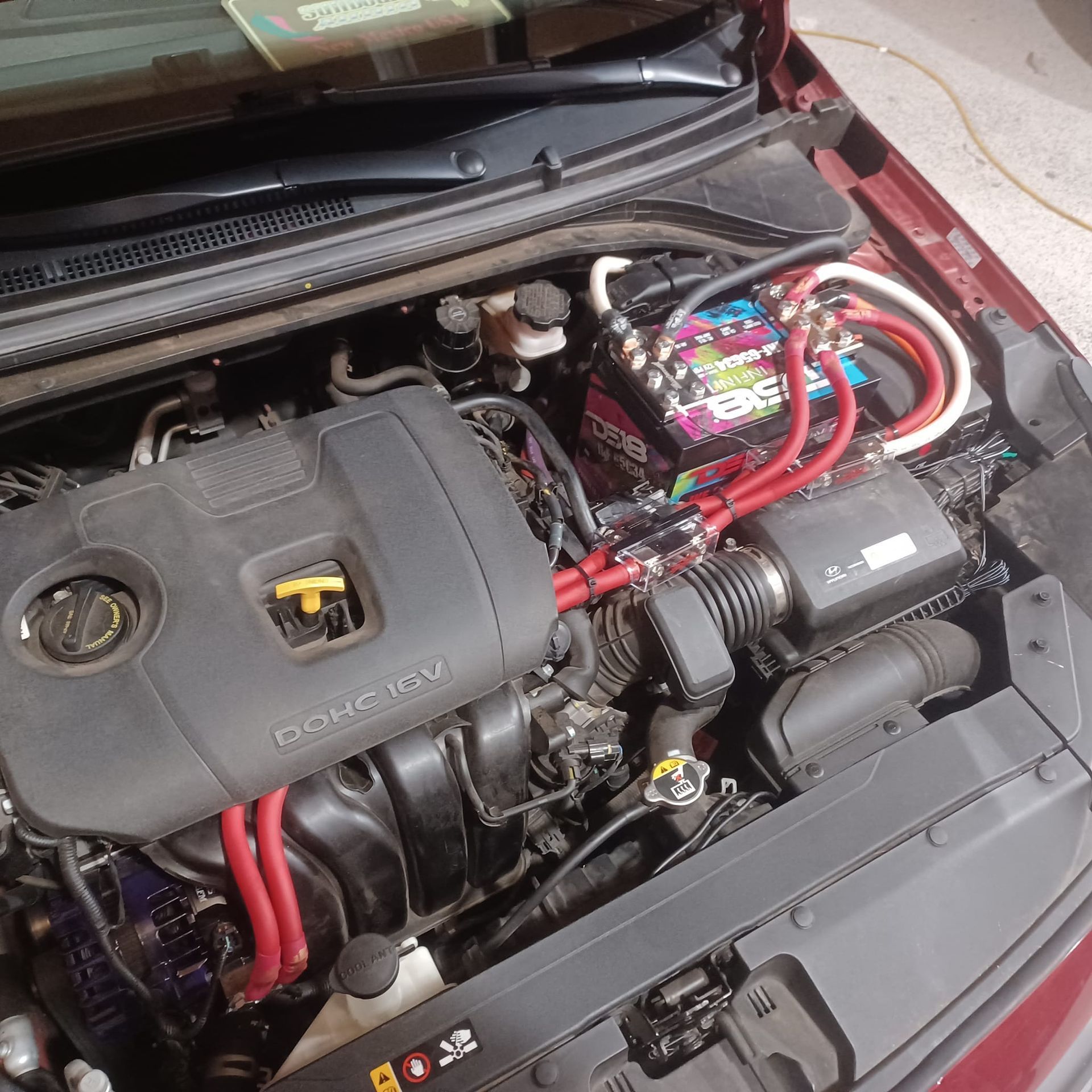 A car engine bay with upgraded wiring for audio. Red and white cables connect to a second battery.