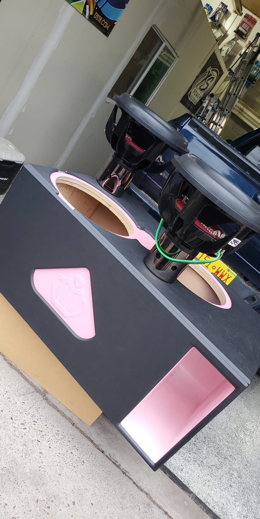 A black speaker box with two subwoofers. The box has pink and tan accents. It sits outside next to a car.