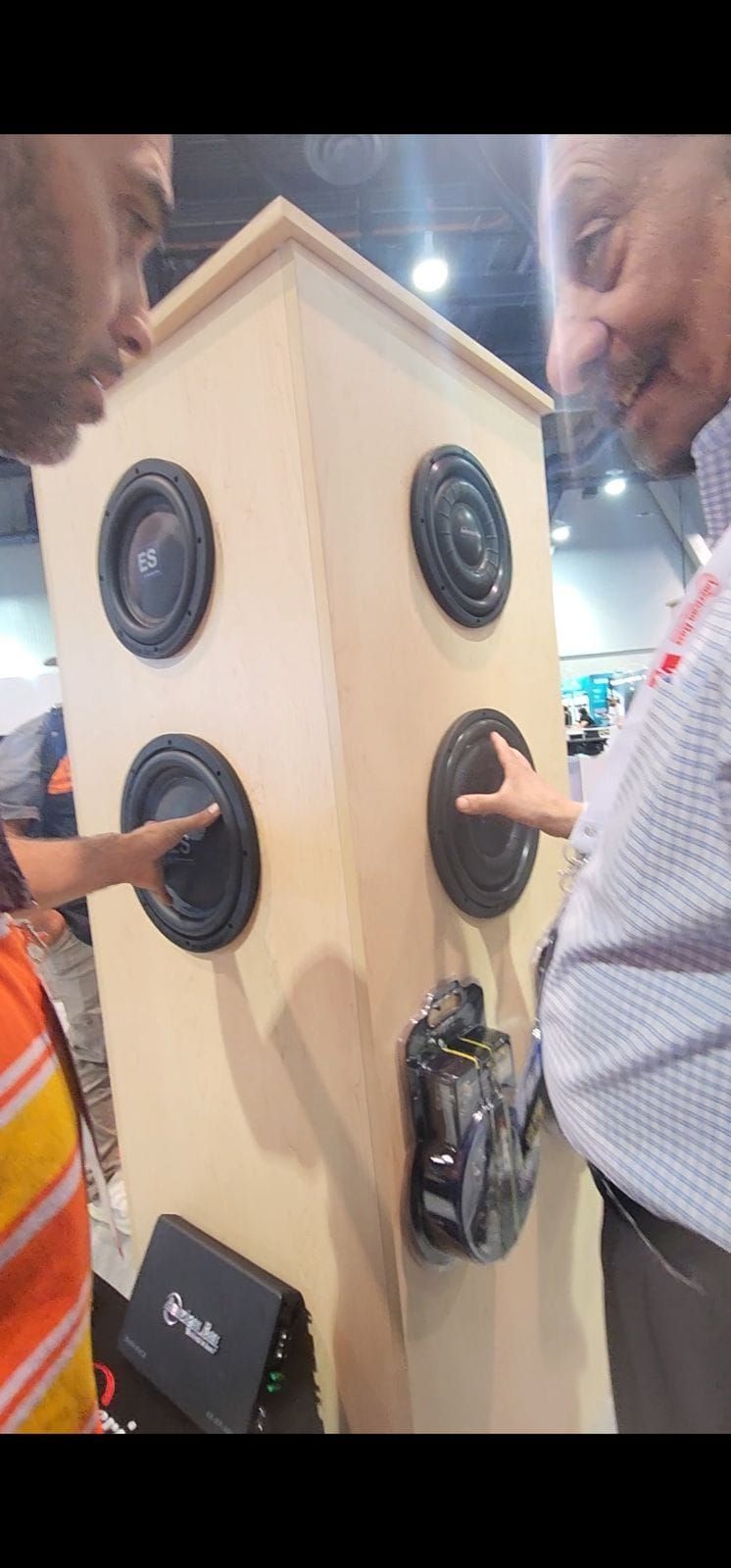 Two men touch the speakers on a tall wooden enclosure. The speakers are arranged on four sides. 