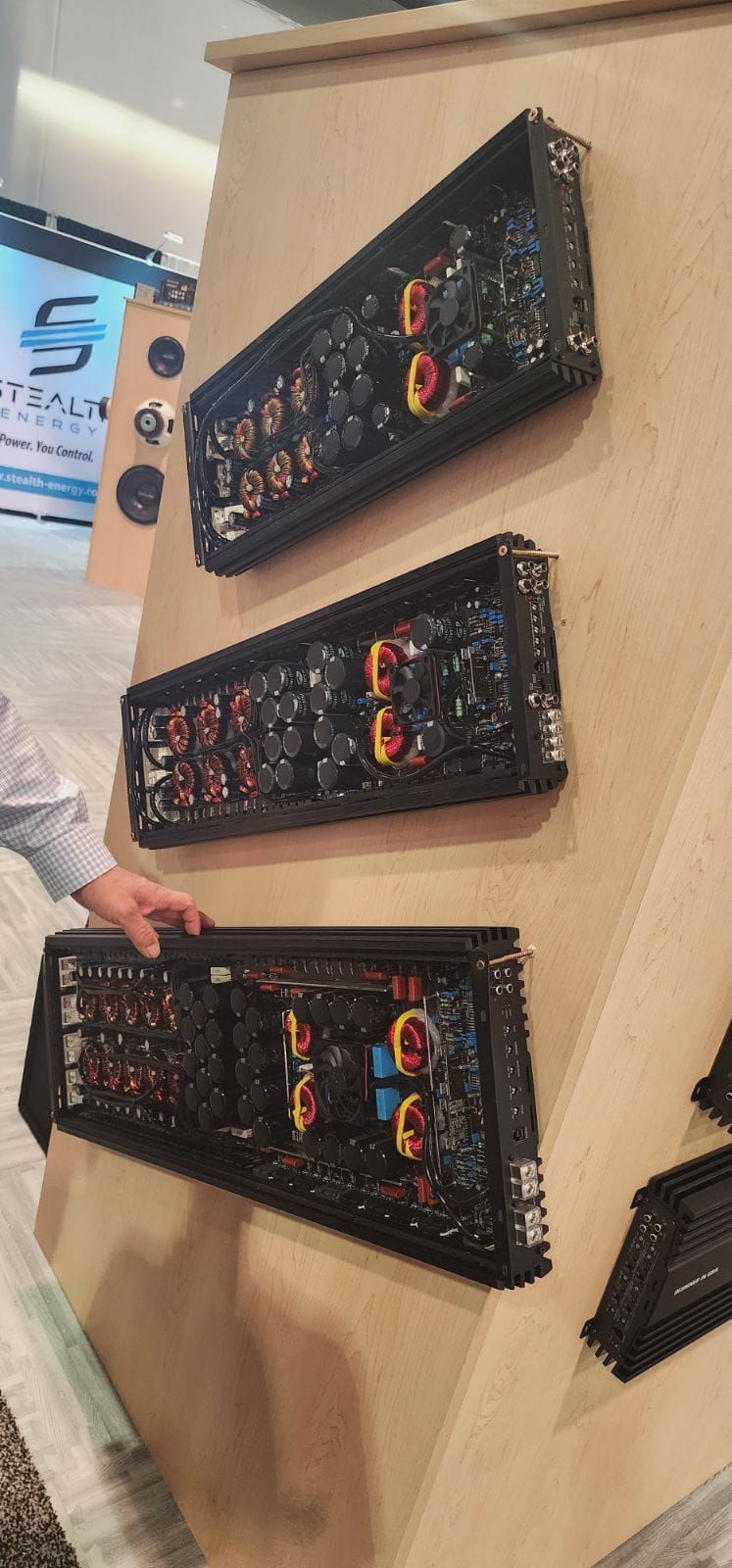 Three black electronic devices are mounted on a light brown angled display. A person's hand is seen near the bottom device.