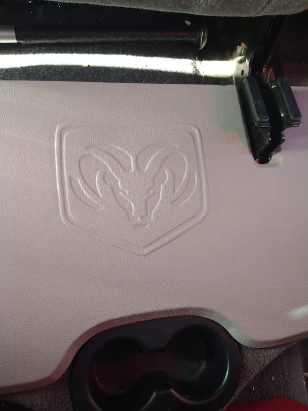 Close-up of a white Dodge Ram logo embossed on a gray surface with cup holders. A black bar is visible above.