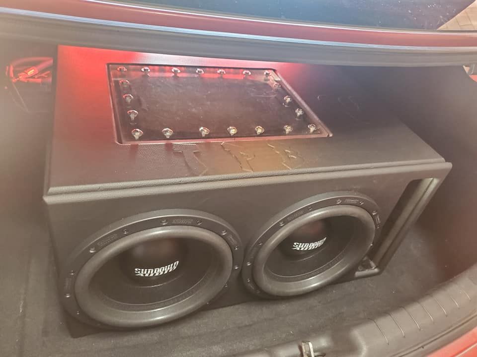 Two subwoofers installed in a black speaker box within a car's trunk. The box is black with a glass-covered top and bolts.