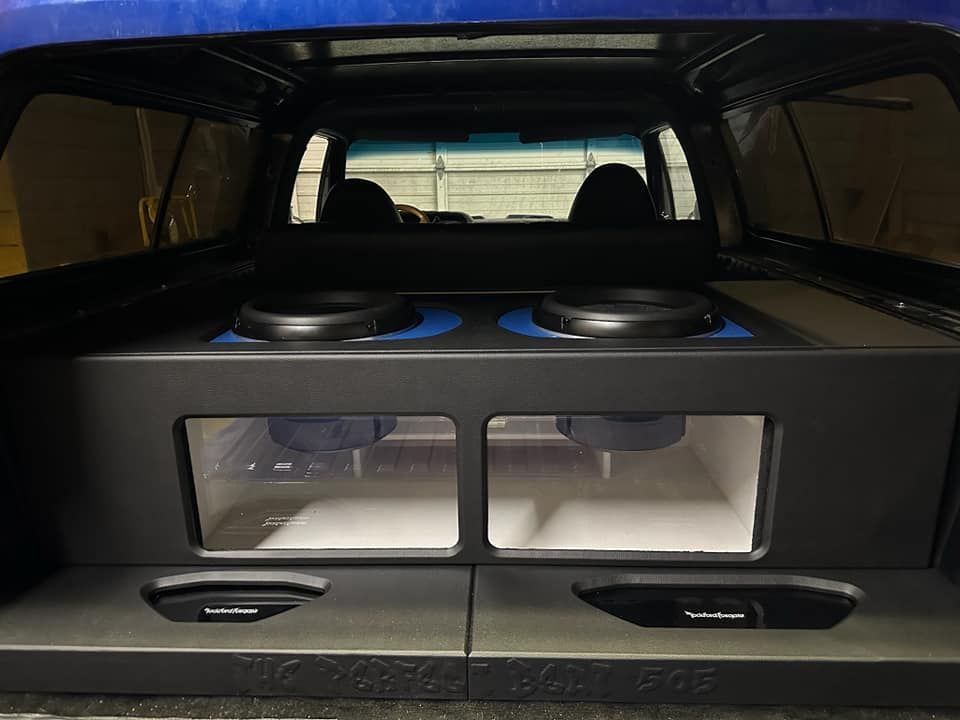 Inside of a blue pickup truck: custom subwoofer setup with two top-facing subs and two visible through a plexiglass window.