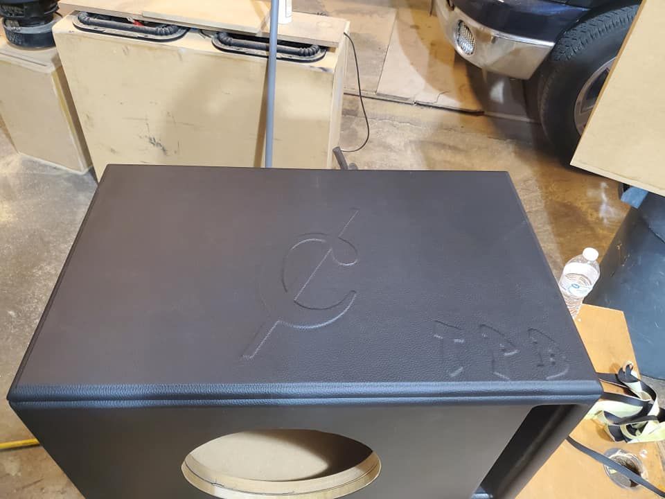 Black subwoofer box with a logo etched on top, a circular cutout, and a garage setting.