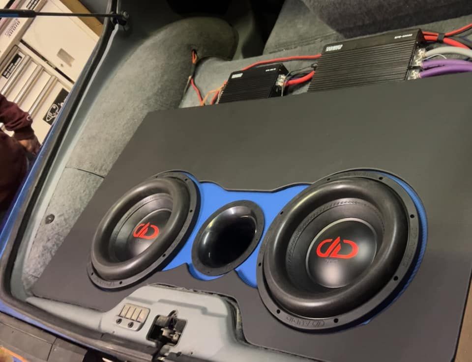 Car trunk with two large subwoofers and a port, mounted on a blue and black board. Electrical wiring and amplifier visible.