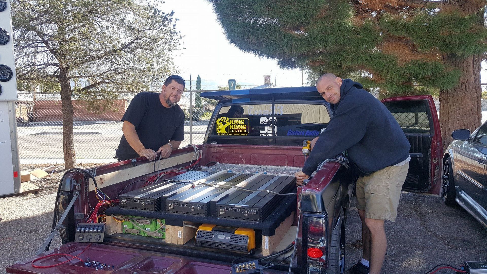 Two men load equipment into the bed of a maroon pickup truck outdoors. One smiles; the other works on gear.