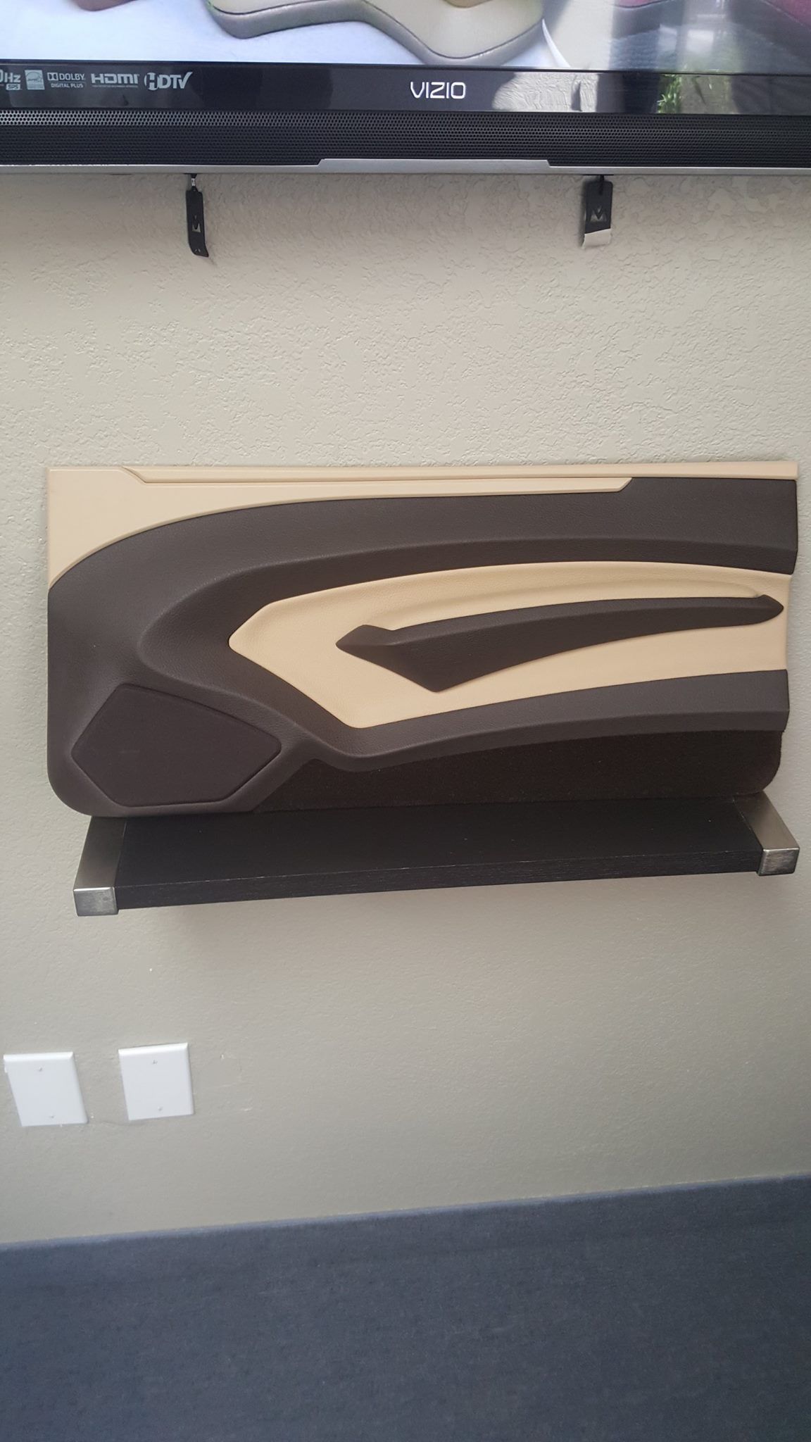 A tan and brown car door panel mounted on a light gray wall beneath a television. Two white electrical outlets are below.
