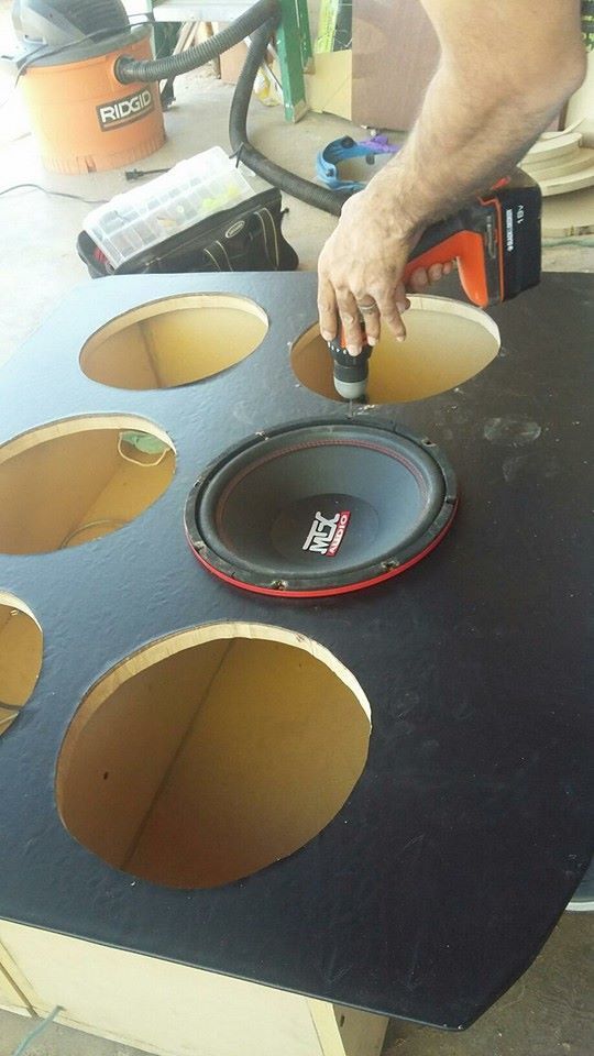 A person installing a subwoofer into a black speaker box, using a power tool. The box has multiple circular openings.