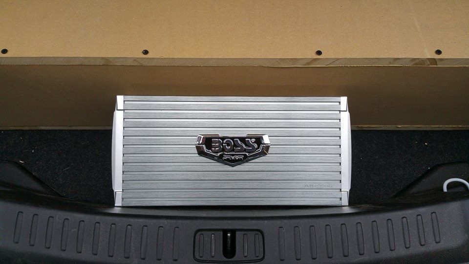 Silver Boss audio amplifier installed in a car trunk.  The amplifier sits on a black mat beneath a wood panel.