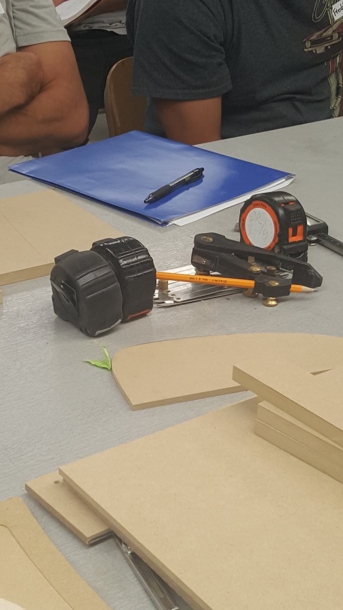 A table with wooden panels, a tape measure, and a blue binder. People are seated at the table.