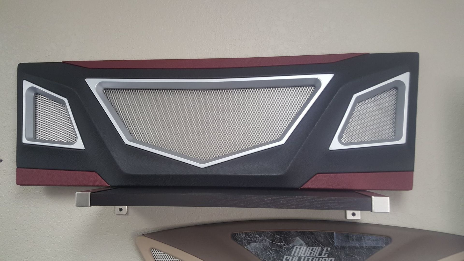 Black, gray, and red custom car dashboard piece with a central mesh grill and side vents, mounted on a wall.