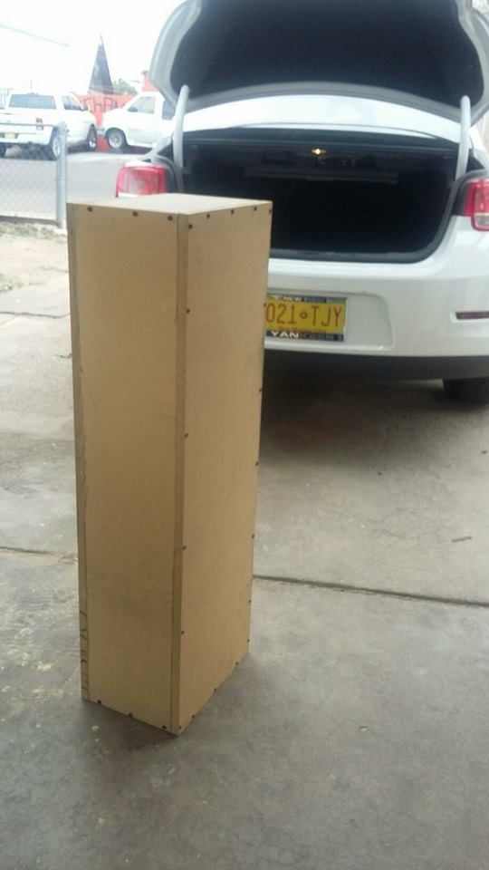 A tall, rectangular brown box sits on a concrete surface in front of a white car with an open trunk.