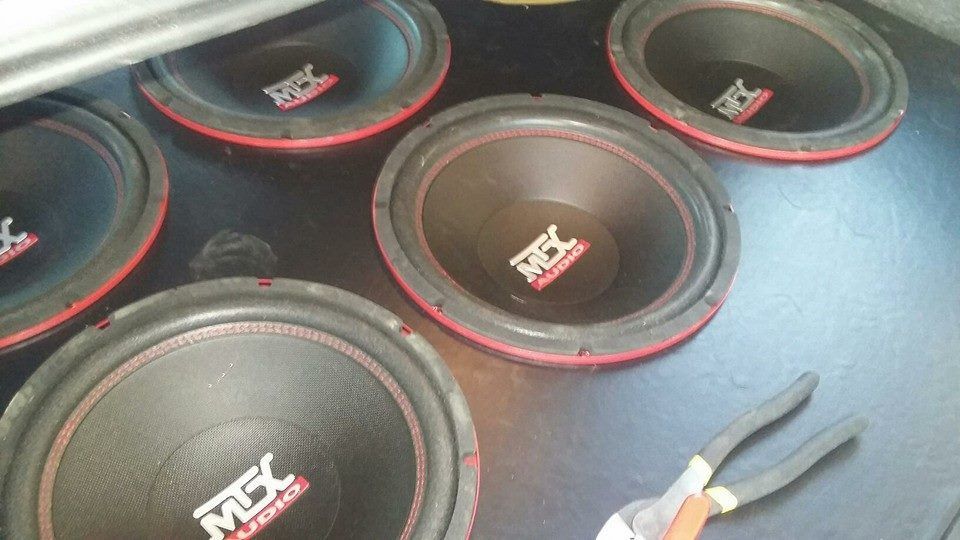 Six car subwoofers with red trim are installed in a vehicle's trunk; pliers rest nearby.