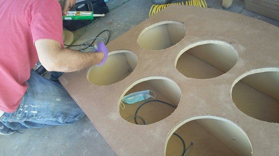 Person sanding a large wood panel with multiple circular cutouts, possibly for speakers.