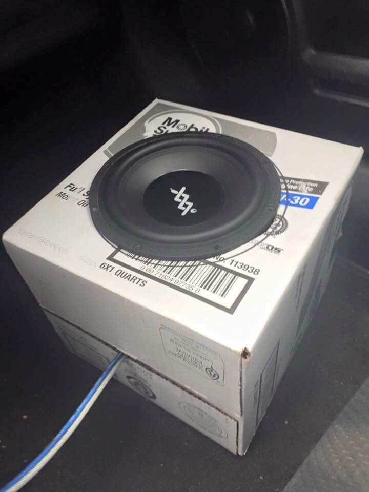 Black subwoofer mounted on a white cardboard box inside a vehicle. Blue and white wire extends from the box.