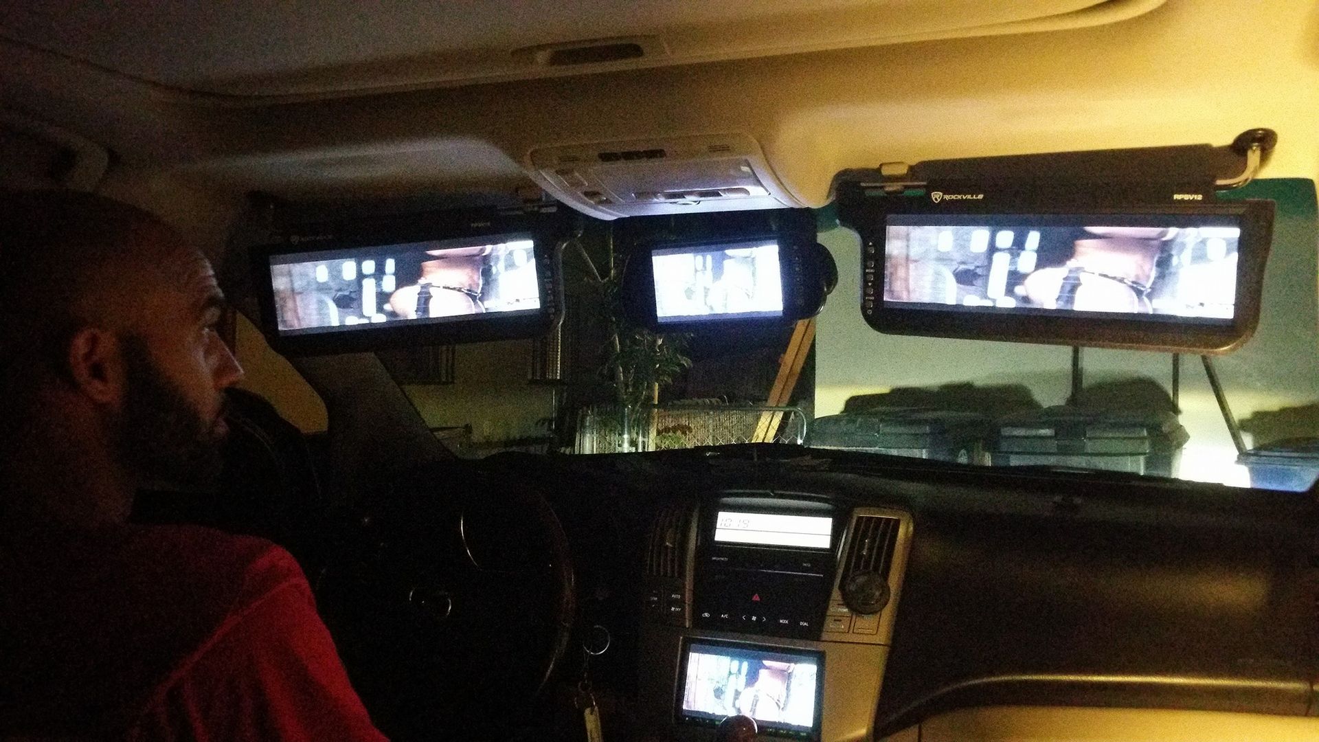 A person sits in a car looking towards three mounted video screens. The screens show a video, with the dashboard visible.