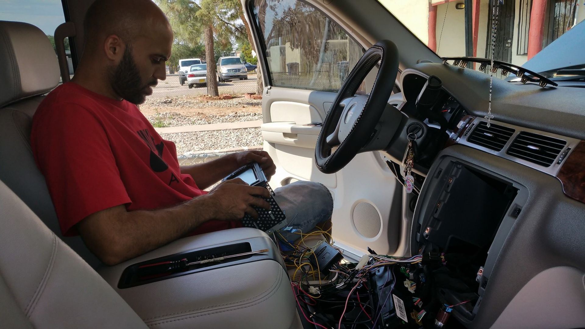A man in a red shirt works on the dashboard of a car, using a tablet. The car's interior is disassembled, with wires exposed.