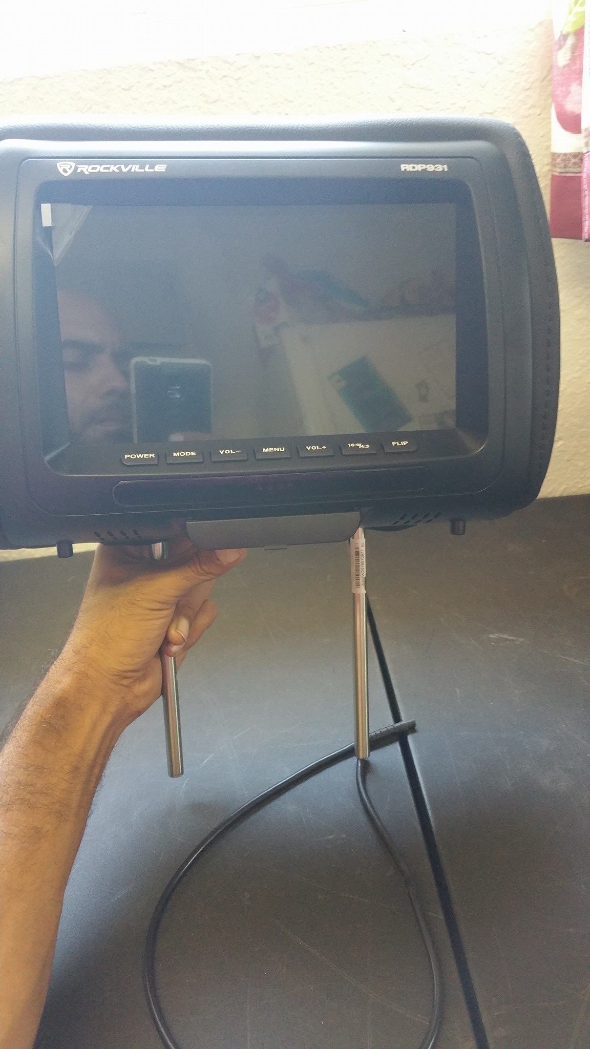 A person holding a black car headrest with an integrated LCD screen, displaying reflections, and metal support rods.