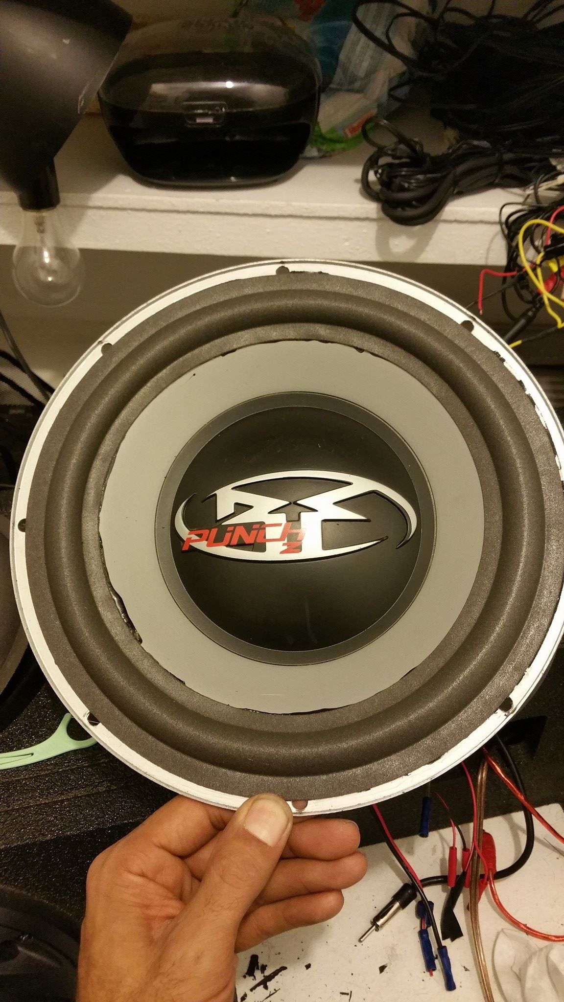 A close-up of a car subwoofer with a gray cone, black dust cap, and white frame. A hand holds the subwoofer at the bottom.