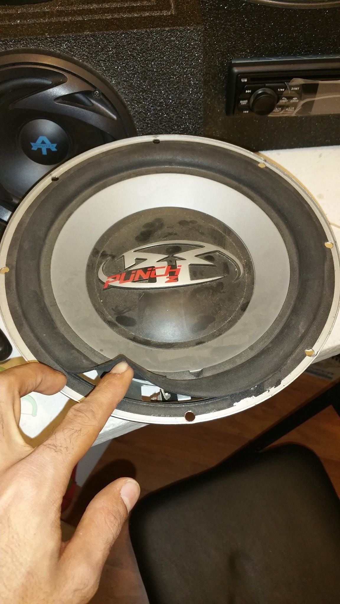 A person's finger points to a detached rubber surround on a white subwoofer speaker.
