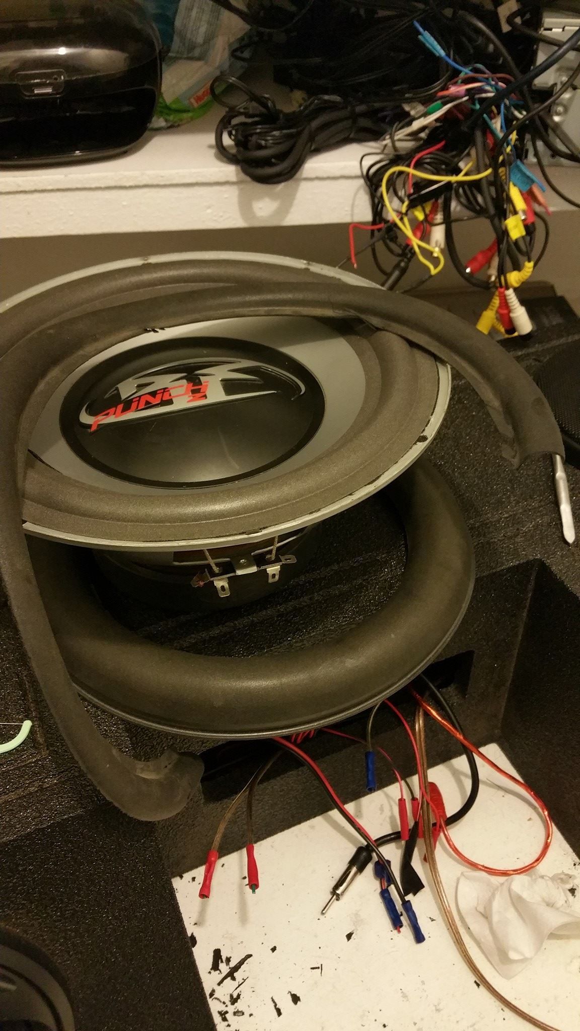 A subwoofer sits on a black surface with wires attached. Foam tubing encircles the speaker. 