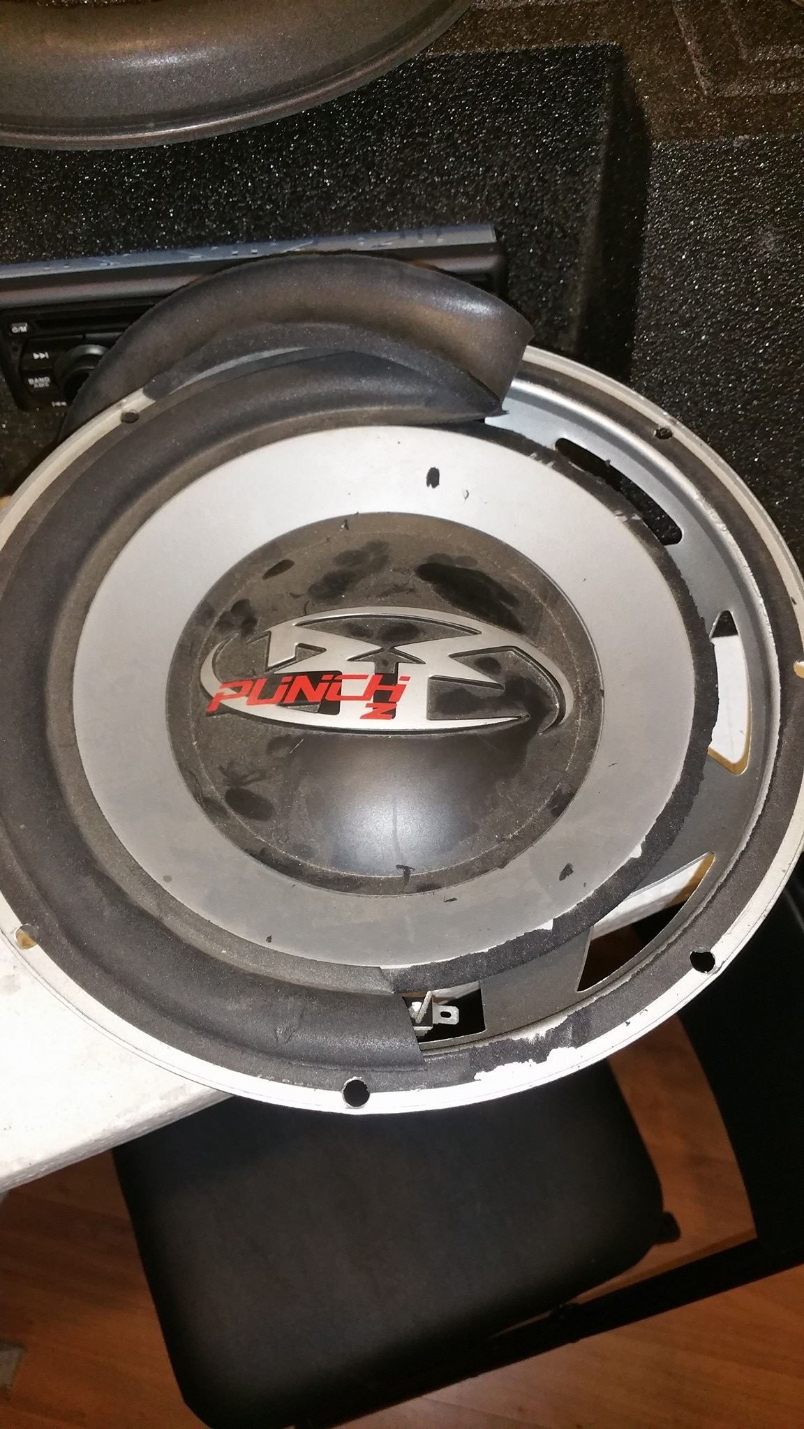 A car subwoofer speaker, with a damaged surround, sits on a black surface. The speaker has a silver frame and a black cone.