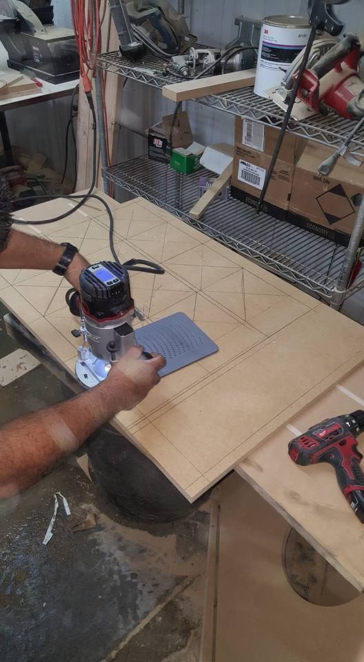 A person uses a router to carve a design into a large wooden board. Workshop setting, with tools and materials visible.
