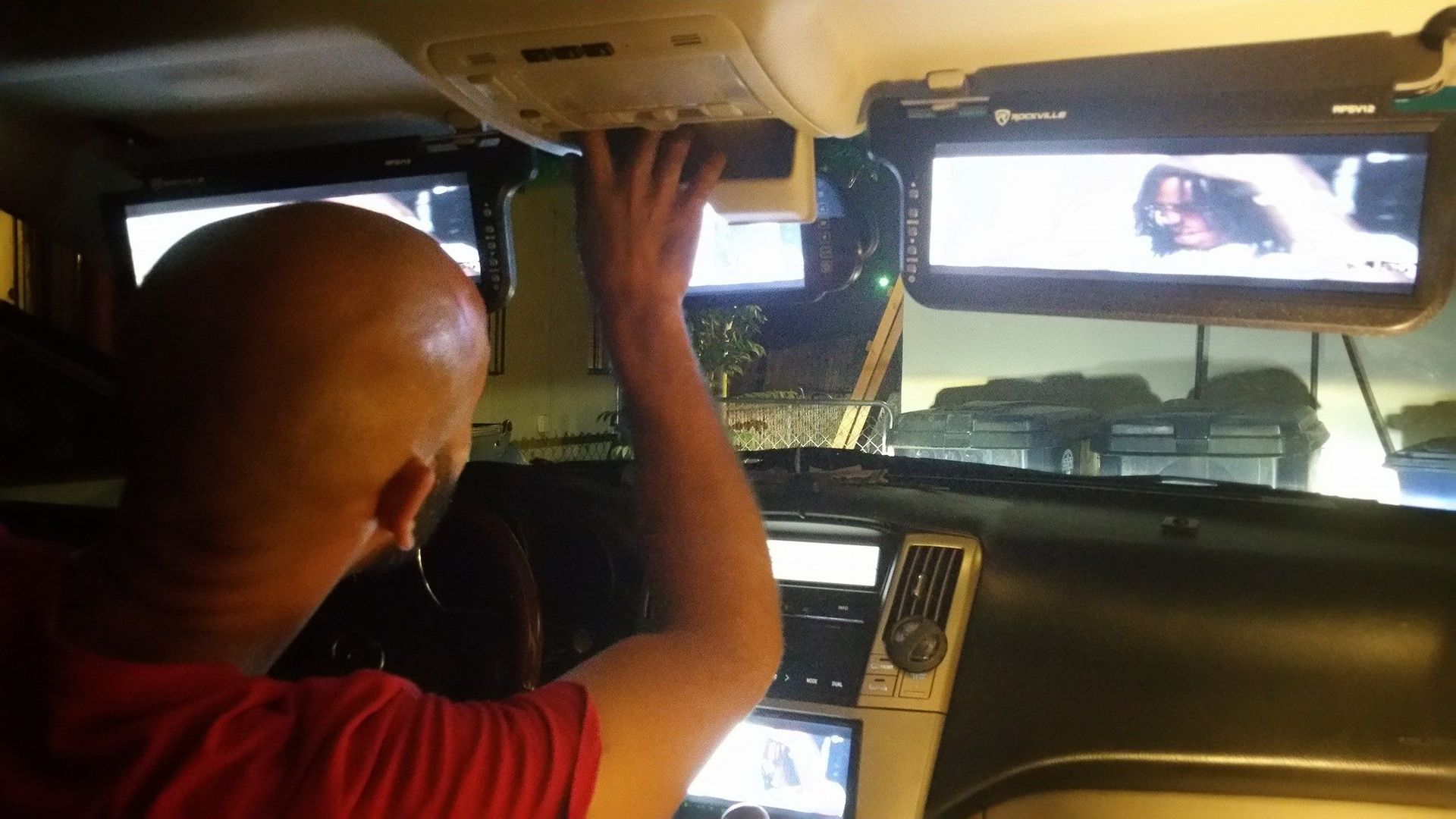 Man in red shirt inside a car, reaching up towards the ceiling with two screens showing video and a control panel.