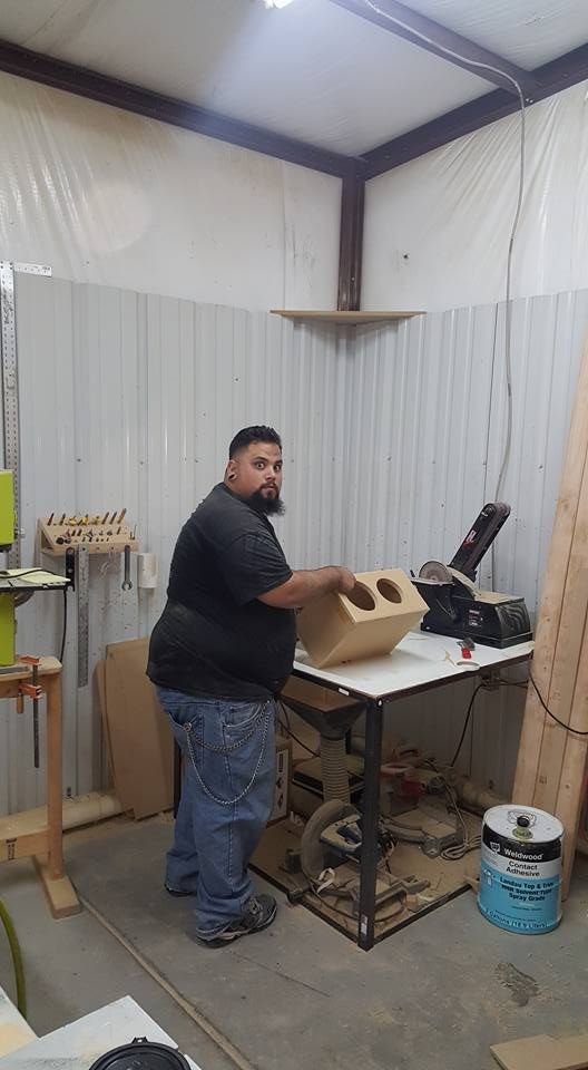 A man works in a woodshop, holding a speaker box. He's wearing blue jeans and a black shirt.